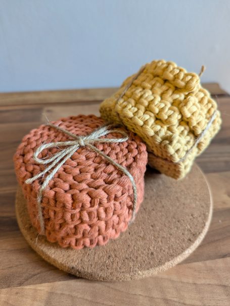 Two decorative, woven coaster packs in orange and yellow on a cork mat.