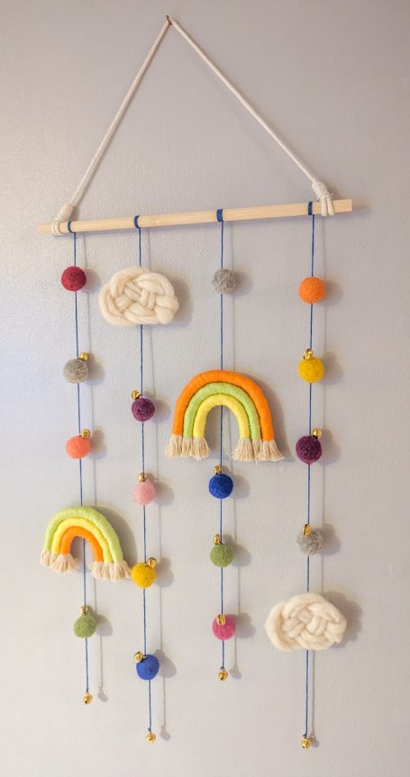 Wall hanging featuring colourful rainbows and clouds with decorative pom-poms.