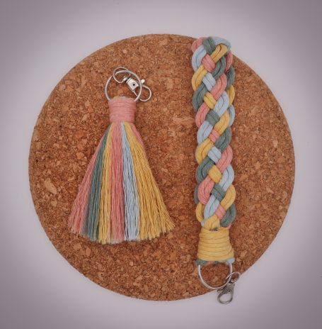 Two handmade keychains: a tassel and a braided cord in pastel colours on a cork surface.
