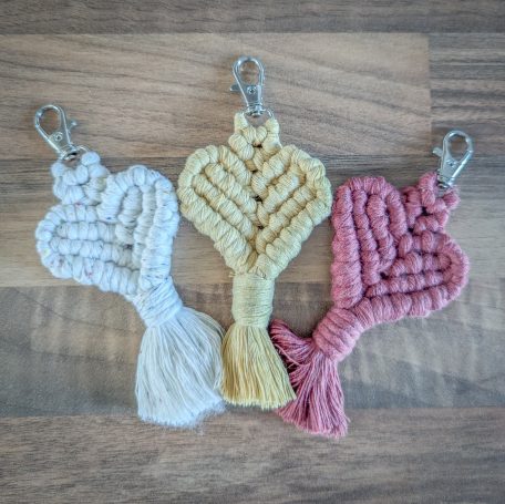 Three macramé heart keychain charms in pastel colours: white, yellow, and pink, with clips.