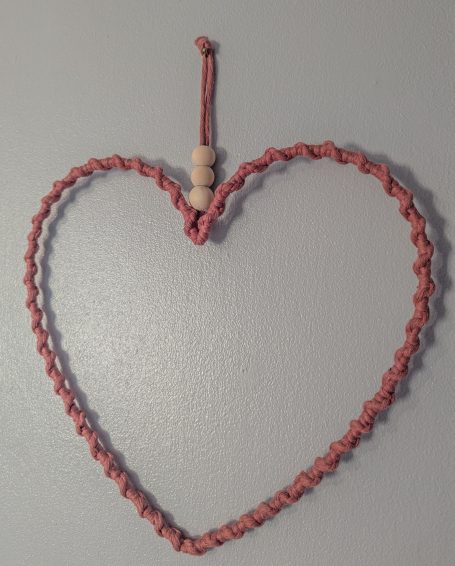 A handwoven heart-shaped decor in pink, accented with a wooden bead.