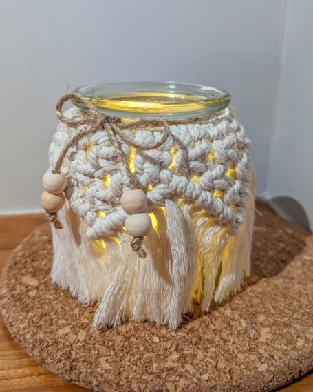 Glass candle holder with macramé detail and wooden beads, emitting warm light.