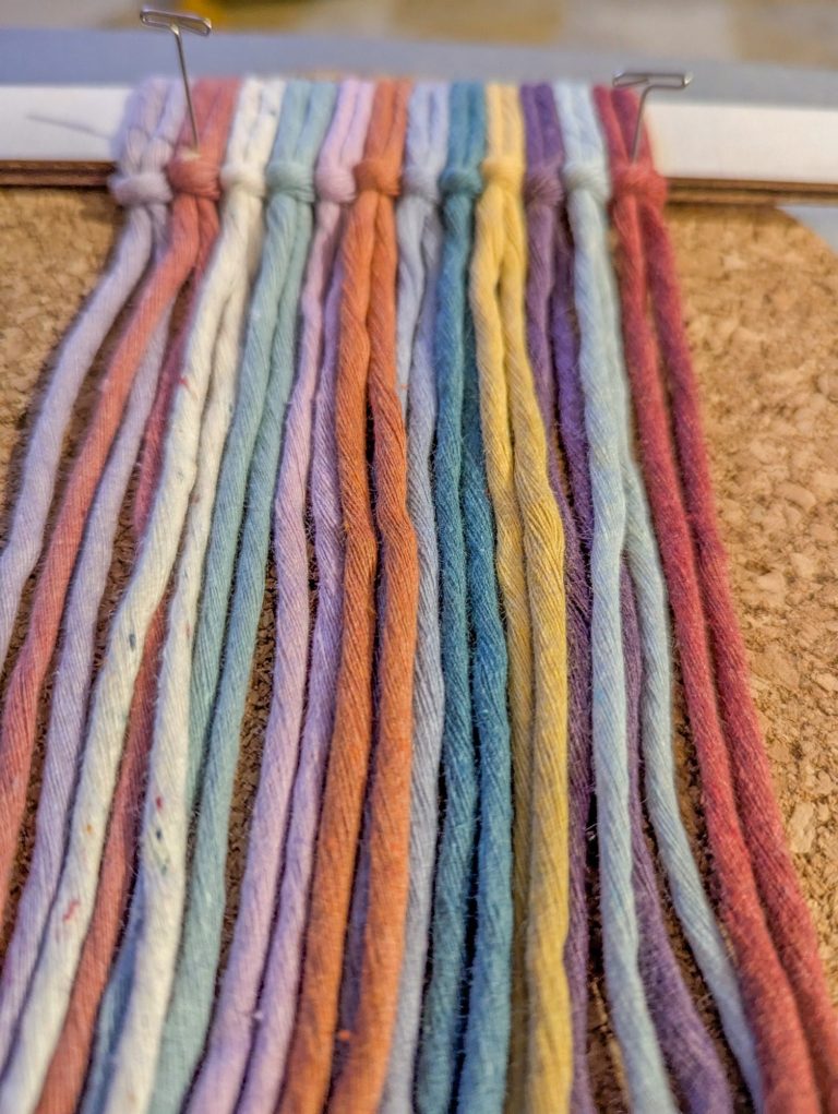 A series of colourful yarn strands arranged vertically on a cork surface.