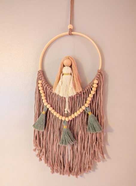 Macramé wall hanging featuring a doll and decorative tassels.