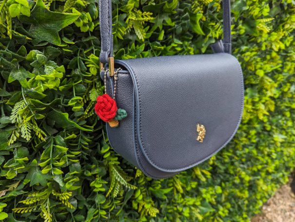 A navy handbag with a red rose bag charm embellishment, hanging on a green hedge.