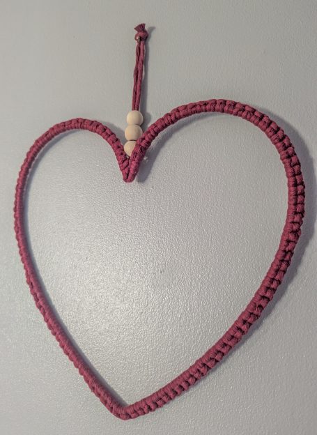 A red woven heart shape with a wooden bead, hanging on a wall.