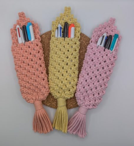 Three macramé pen holder pouches in peach, yellow, and pink, filled with writing instruments.
