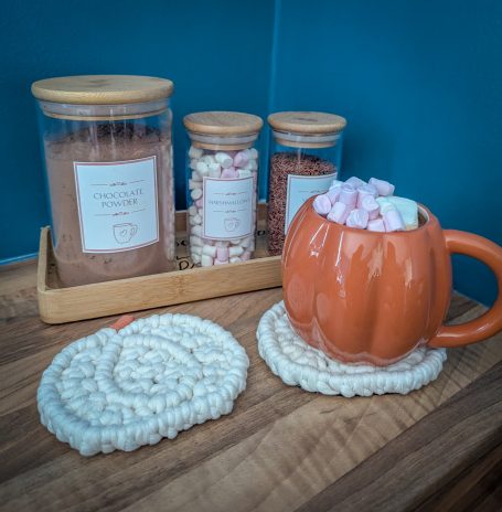 A pumpkin-shaped mug filled with marshmallows, beside jars of hot chocolate mix sitting on macramé pumpkin coasters.
